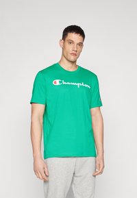 Vihreä lyhythihainen T-paita, joka on valmistettu puuvillasta. Tässä paidassa on suuri valkoinen "Champion"-logo rinnassa ja pieni logo hihassa.