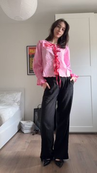 Jeune femme debout dans une chambre, portant un chemisier rose avec une broderie rouge et un pantalon large noir, les mains dans les poches.