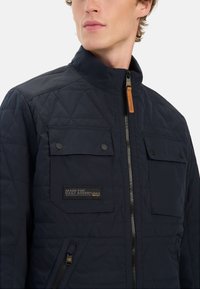 Granatowa pikowana kurtka z stójką, z przednim zamkiem błyskawicznym, dwoma kieszeniami na klatce piersiowej z zapięciem na zatrzask oraz metką z napisem "STWORZONE DO CODZIENNYCH PRZYGÓD".