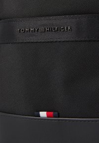 Black textile bag featuring a smooth leather base and a stitched red, white, and blue accent. Contains embossed "TOMMY HILFIGER" logo detail.