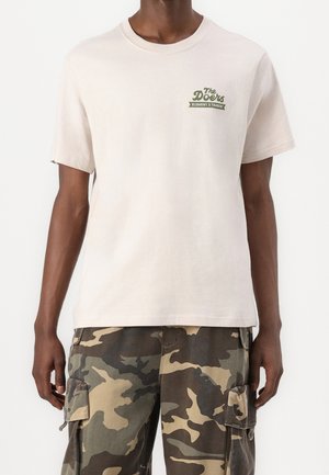 Homme portant un t-shirt beige clair avec le texte "The Doers Element x Timber" sur la poitrine et un pantalon cargo camouflage vert et marron.