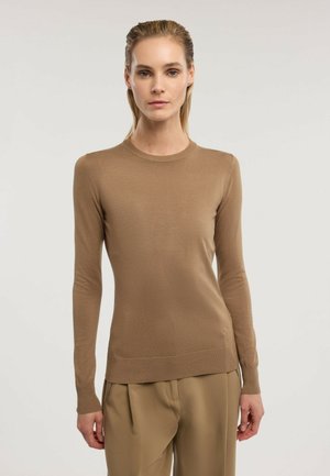 Kobieta o blond włosach, ubrana w brązowy sweter z długim rękawem i beżowe spodnie, stojąca na jednolitym jasnym tle.