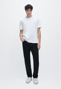 Bílé tričko s krátkým rukávem a kulatým výstřihem, kombinované s černými kalhotami a šedými teniskami. Jednoduchý design, lehká látka a neutrální barvy.