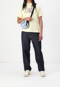 Osoba ubrana w żółtą koszulkę z grafiką, ciemnoniebieskie dżinsy, beżowe sneakersy i jasnoniebieską torbę na ramię, na tle jednolitego białego tła.