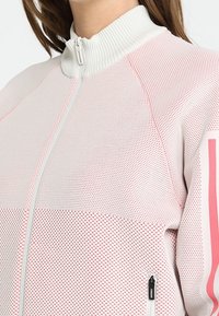 Chaqueta blanca con cremallera y un diseño de malla texturizada, con acentos en rosa y un cuello alto. Incluye cremallera y bolsillos laterales.