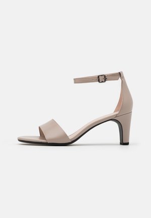 Beige sandal med åben tå, lav hæl og ankelrem med en lille sort spænde, vist på en hvid baggrund.