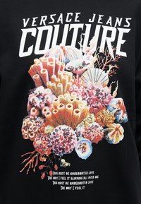 Texto "Versace Jeans Couture" sobre un diseño colorido de arrecife de coral y conchas marinas con pequeñas lentejuelas sobre fondo de tela negra.