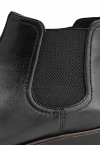 Botte cheville en cuir noir avec panneau latéral élastique, finition texturée, couture visible et bord de semelle marron.