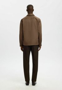 Homme debout, dos à la caméra, portant une chemise à manches longues marron, un pantalon marron foncé et des chaussures habillées noires sur fond blanc.