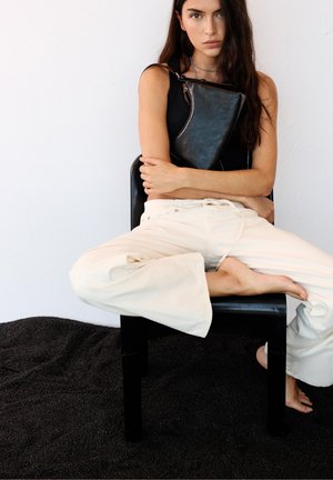 Frau mit langen dunklen Haaren sitzt im Schneidersitz auf einem schwarzen Stuhl, trägt ein schwarzes Tanktop, cremefarbene Hosen und hält eine schwarze Ledertasche vor der Brust.