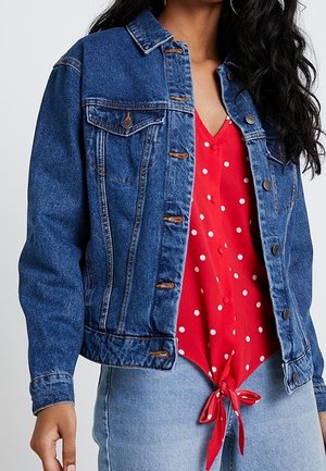 Woman wearing blue denim jacket over red polka dot blouse tied at waist and light blue jeans.
