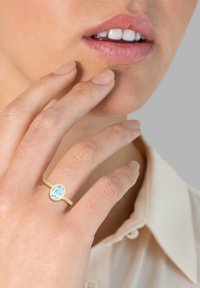 Bague en or avec une gemme ovale bleu clair, montée sur un bandeau fin. La main repose près du visage avec du vernis à ongles naturel.
