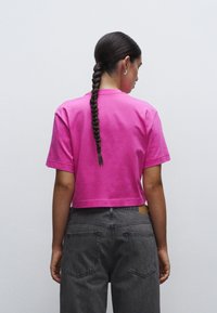 Zkrácený tričko v živé růžové bavlně, s krátkými rukávy a kulatým výstřihem. V kombinaci s šedými džíny s vysokým pasem a zadními kapsami.