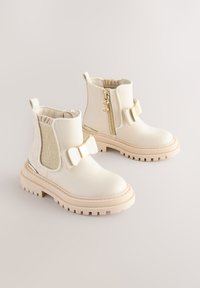 Bottes blanches en cuir à la cheville avec un nœud décoratif, un panneau latéral élastique, une fermeture éclair et une semelle en caoutchouc épaisse avec un motif texturé.