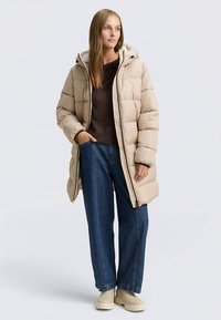 Bėrino spalvos pūkinė striukė su gaubtu, su siuvinėta danga, priekyje užtrauktukas ir šoninės kišenės, derinama su mėlynais plačiomis kojų kelnėmis ir bėrino spalvos avalyne.