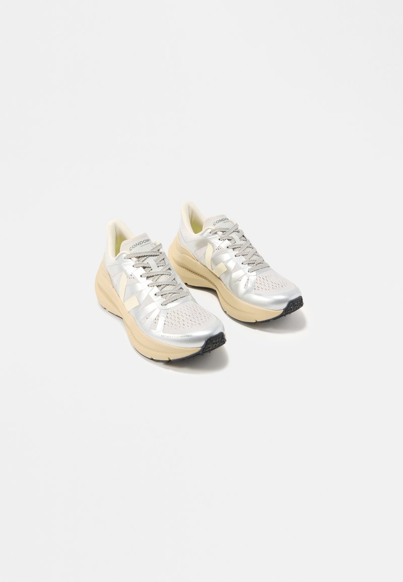 Ζευγάρι ασημένιων και μπεζ sneakers με κορδόνια και παχιά σόλα, με την επωνυμία «Condor», εκτεθειμένα σε λευκό φόντο.