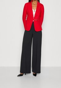 Rød blazer med skræddersyet design, der har en dekorativ guldkædeaccent ved taljen, parret med sorte brede bukser.