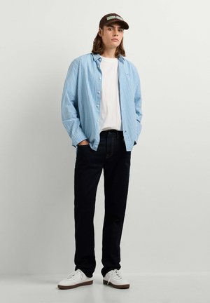 Junger Mann trägt ein blau kariertes Hemd über einem weißen T-Shirt, schwarze Jeans, weiße Sneakers und eine dunkle Kappe, steht mit den Händen in den Taschen.