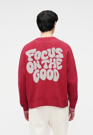 Άτομο που φοράει κόκκινο πουλόβερ με άσπρο κείμενο "Focus on the Good" στην πλάτη, συνδυασμένο με ανοιχτόχρωμο παντελόνι, γυρισμένο πλάτη στην κάμερα.