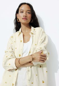 Chemise jaune clair à boutons avec broderie florale et manches retroussées. Débardeur blanc en dessous. Bracelets en or au poignet.