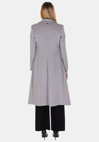 Cappotto lungo grigio con collo appuntito, silhouette strutturata e pieghe posteriori. Abbinato a pantaloni neri a gamba larga e scarpe con tacco.