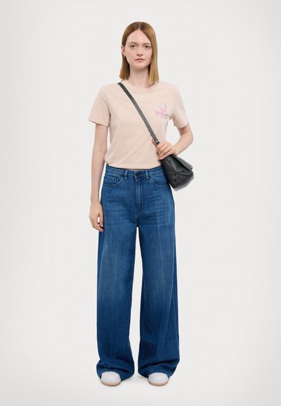 Beige bavlněné tričko s růžovým potiskem králíka, v kombinaci s volnými modrými džíny. Modelka drží černou crossbody kabelku. Viditelné bílé tenisky.