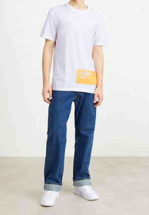 Weißes T-Shirt mit kurzen Ärmeln und orangefarbenem Grafikdruck am Saum, kombiniert mit blauen Jeans, die an den Bündchen hochgekrempelt sind, und weißen Turnschuhen.