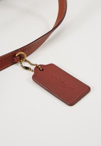 Cintura in pelle di un marrone intenso con accessori in oro. L'etichetta attaccata presenta un logo impresso ed è fissata con una catena di perline. Forma rettangolare.
