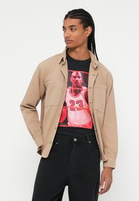 En tan knap-op skjorte over en sort grafisk t-shirt med et rødt og sort basketball design, parret med sorte jeans.
