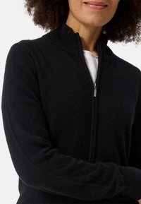 Mustat vetoketjuhuppari, jossa on korkea ribbikaarroke, valmistettu pehmeästä materiaalista. Ominaisuuksiin kuuluu pitkät hihat ja sileä pinta. Huomattava vetoketj yksityiskohtana.