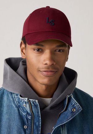 Casquette de baseball rouge avec logo "LS" brodé en marine ; portée avec un sweat à capuche gris et une veste en denim, affichant une coupe classique et des détails déchirés.