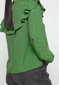 Green and black gingham blouse with ruffle detailing along the shoulders, a round neckline, and a visible zipper at the back.