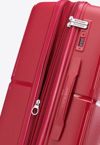 Maleta rígida roja con superficie texturizada, doble cremallera, candado de combinación y tirador de cremallera con marca. Cuenta con un asa resistente.