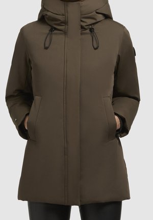 Femme portant un manteau d'hiver matelassé vert olive longueur genou avec capuche et mains dans les poches avant.