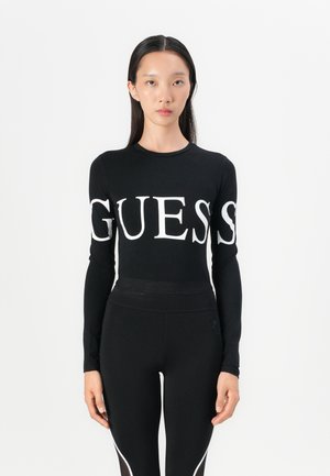 Maglia nera a maniche lunghe con grande logo bianco "GUESS" sul petto, abbinata a leggings neri dal design elegante e con dettagli bianchi.