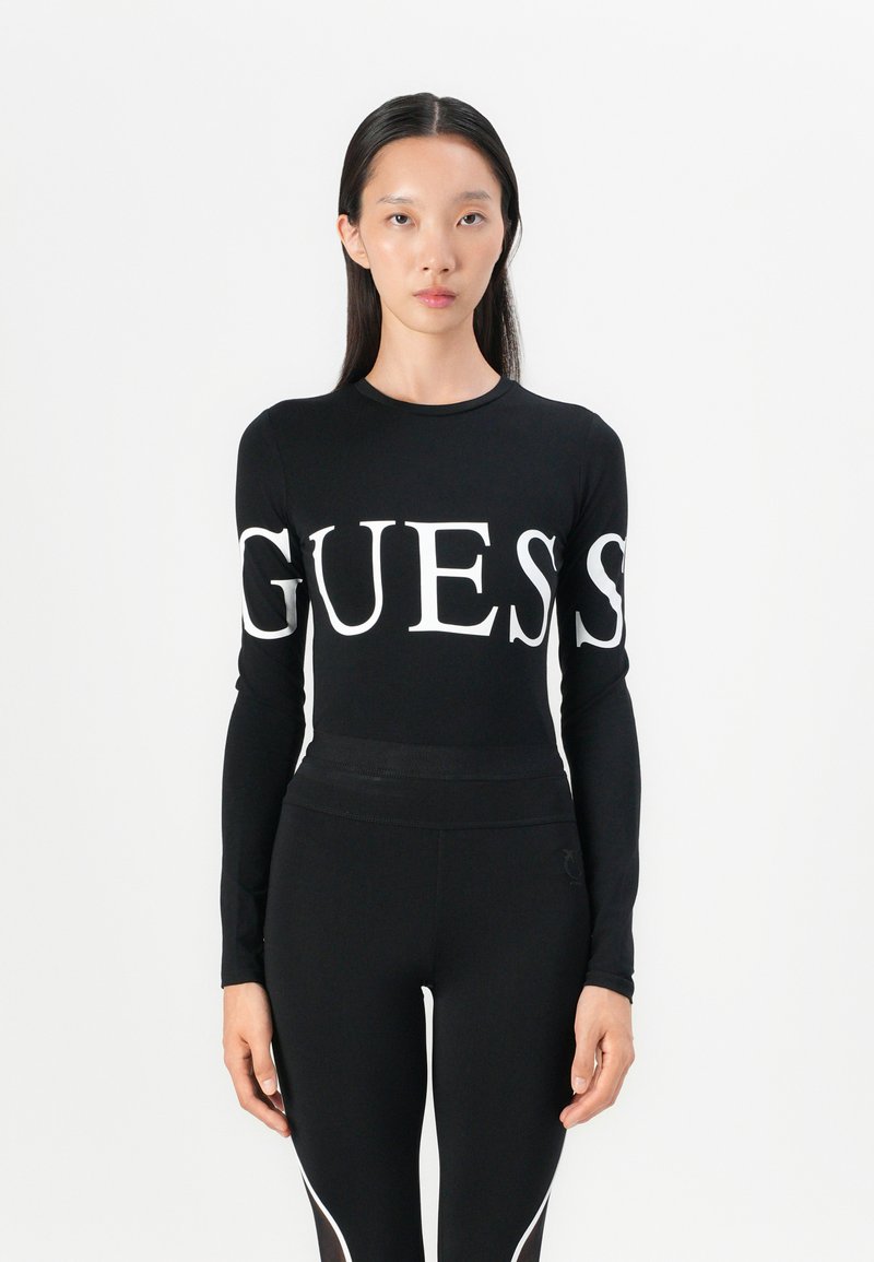 Top noir à manches longues avec un grand logo blanc "GUESS" sur la poitrine, associé à des leggings noirs au design épuré avec des accents blancs.