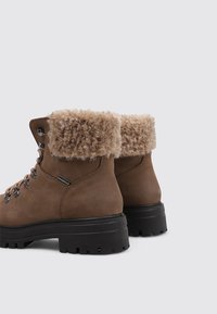 Braune Leder-Wanderschuhe mit einem strukturierten beigen Kunstfellbund, Schnürverschluss und robusten schwarzen Gummisohlen. Mit Ösen für die Schnürsenkel.