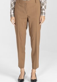 Brązowe spodnie na miarę o prostym kroju, z przednimi zakładkami i gładką fakturą. Noszone z wzorzystą koszulą z długim rękawem i czarnymi szpilkami.