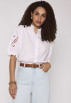 Donna con capelli ricci che indossa una camicetta bianca con inserti in pizzo, jeans azzurri a vita alta, cintura marrone e gioielli dorati, in posa.