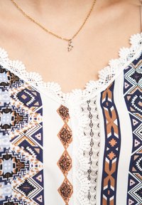 Blusa con patrón de diseños geométricos en navy, blanco y marrón, con un borde decorativo de encaje a lo largo del escote. Collar dorado visible.
