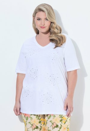 Blonde Frau mit welligem Haar trägt ein weißes Shirt mit V-Ausschnitt und kleinen Strassstein-Clustern sowie geblümte Hose, steht vor weißer Wand.