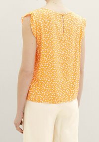 Pomarańczowa bluzka w kwiatowy wzór, krótki rękaw i detal w formie otworu w tyle, wykonana z lekkiego materiału. Połączona z kremowymi spodniami.