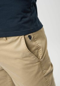 Svijetlosmeđi pamukasti hlače slim fit kroja, s bočnim džepovima i malim crnim izvezenim logotipom na lijevom džepu.