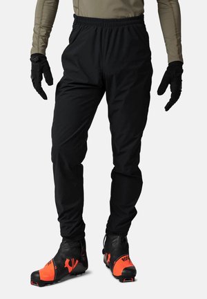 Black, windproof pants with an elastic waistband, paired with black and orange ski boots featuring a logo. Gloves add grip and warmth.