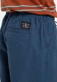 Modré bavlněné šortky s elastickým páskem, bočními kapsami a zadní kapsou, na které je černá textilní nápis s bílým textem.