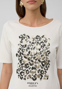 Biały t-shirt z krótkim rękawem, ozdobiony czarnym i złotym wzorem w panterę, z napisem "STUDIO N°1 COLLECTION" na dole.