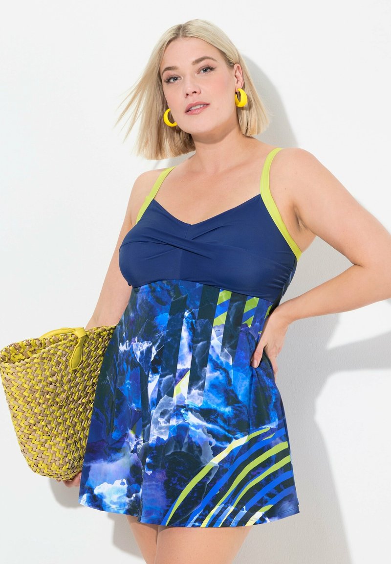 Robe de bain à motifs bleus et verts avec un col en V, dotée d'un haut uni bleu marine et d'une jupe évasée. Accessoirisée d'un sac en osier jaune.