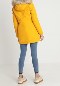 Yellow parka with a fur-lined hood, featuring a mid-length cut and straight sleeves, paired with blue skinny jeans and beige sneakers.