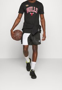 Sort t-shirt med "BULLS" logo, sort og hvide basketball shorts, sorte sneakers med neon detaljer, holder en basketball.
