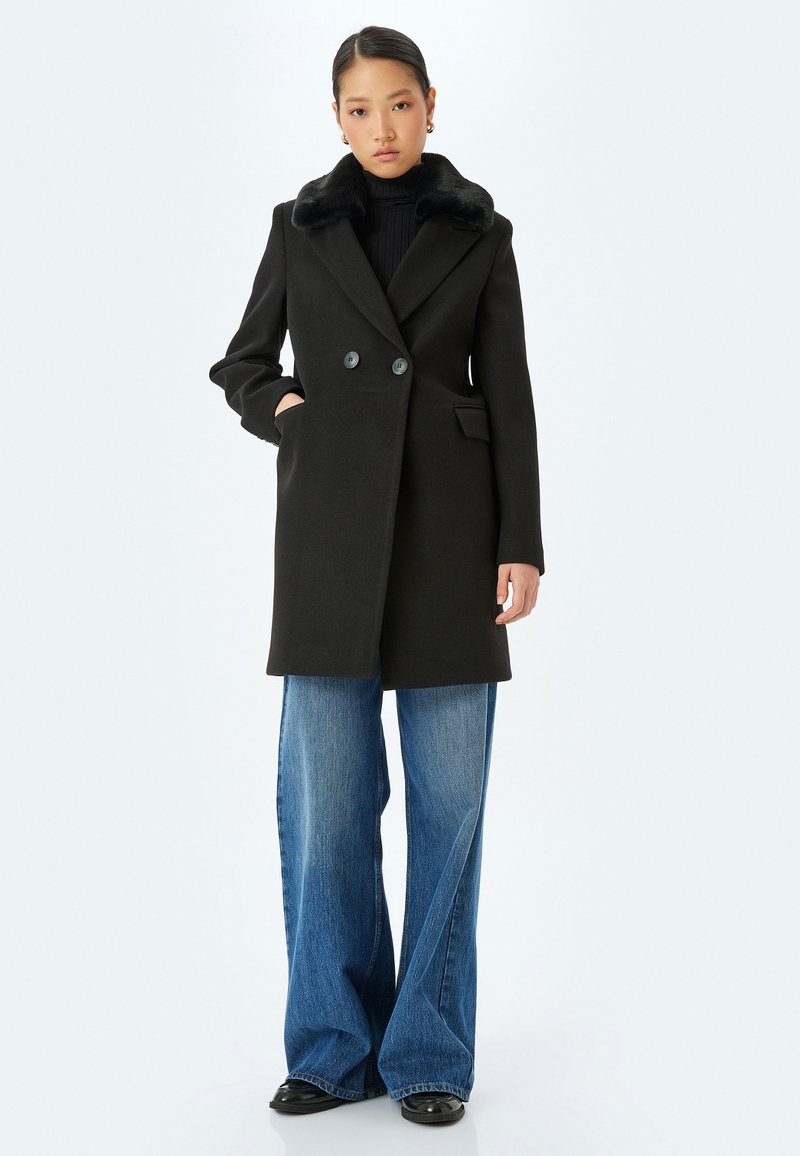 Sort uldfrakke med en krave af falsk pels, dobbeltknaplukning, sidelommer, parret med bredbenede blå denimjeans.